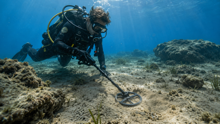 Underwater metal detecting scene with detector coil near rocks and coins.